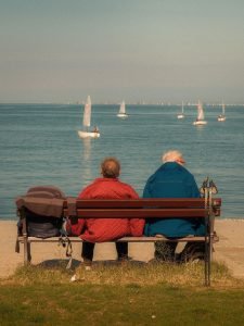 An elderly couple sitting on a bench by the seaside, watching sailboats on a calm day.